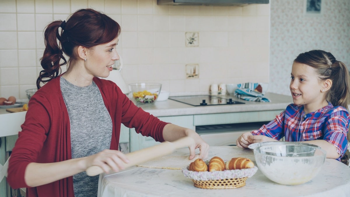 Young mother rolling dough and talking to her little cute daughter while cooking in the kitchen on weekend. Family, food, home and people concept